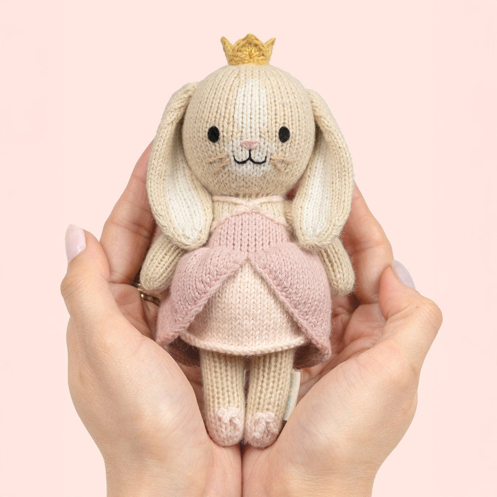 Cuddle + Kind Tiny Rose the Princess, a hand-knit bunny in a pink dress and yellow crown, is gently held in two hands against a soft pink background.