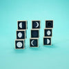 Nine Uncle Goose Moon Phase Blocks, each featuring illustrations of different lunar phases, are arranged in three stacks against a solid turquoise background, capturing the beauty of lunar cycles and evoking the wonder of a possible lunar eclipse.