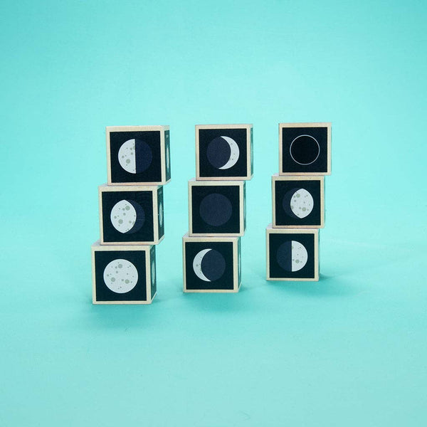 Nine Uncle Goose Moon Phase Blocks, each featuring illustrations of different lunar phases, are arranged in three stacks against a solid turquoise background, capturing the beauty of lunar cycles and evoking the wonder of a possible lunar eclipse.
