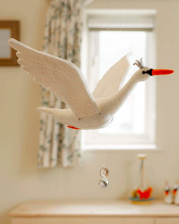 The Handmade Swan Mobile, a plush white swan with a small crown, elegantly flaps as it hangs. Made from organic felt and eco-friendly dyes, it features two shiny bells. Hanging in front of a window, it captures streaming light to softly illuminate the room.