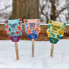 A Pollinator Seed Pop (Single) stands upright in the snow with "Butterfly," "Song Bird," and "Humble Bee" labels, ready to help grow a vibrant pollinator garden. Trees and wood offer a natural backdrop for this colorful wildflower seed pop.