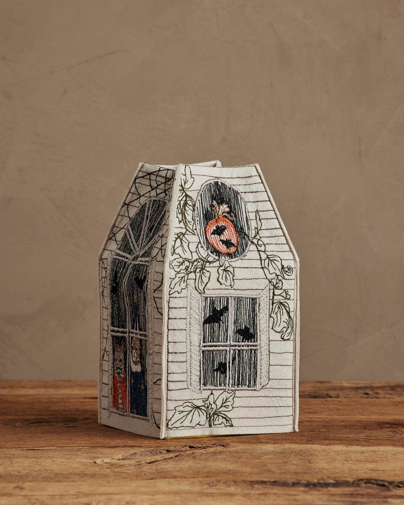The Coral & Tusk Haunted House Tissue Box Cover, featuring jack-o’-lanterns, bats, and cobweb embroidery, sits on a wooden surface against a plain beige background.