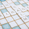 Close-up of a wood-framed Scrabble Bianco Edition board featuring white letter tiles spelling out “MIRAGE” on light blue and gold patterned squares.