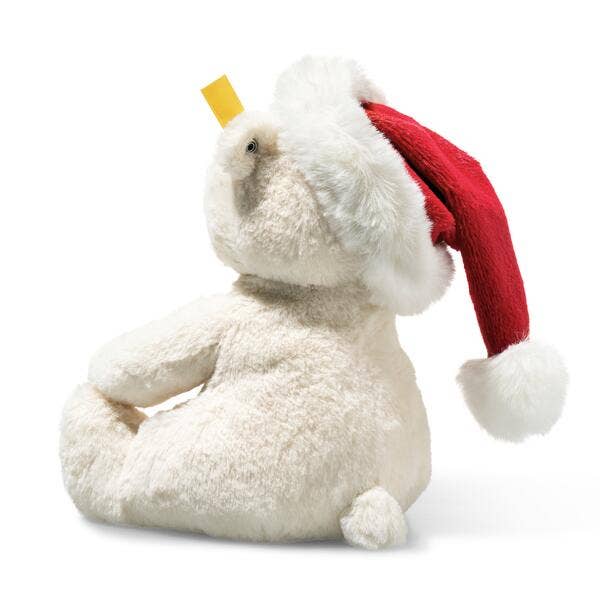 The Steiff Cozy Christmas Rico Polar Bear, a white plush bear in a Santa hat, sits upright on a white background with a yellow tag near its nose, bringing festive charm to the scene.