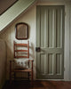 A wooden ladder-back chair with a Coral & Tusk Aspen Log Bunnies Pocket Pillow sits against a cream wall beneath a small mirror, beside a closed muted green paneled door with a dark metal handle.