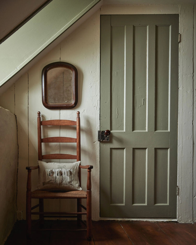 A wooden ladder-back chair with a Coral & Tusk Aspen Log Bunnies Pocket Pillow sits against a cream wall beneath a small mirror, beside a closed muted green paneled door with a dark metal handle.