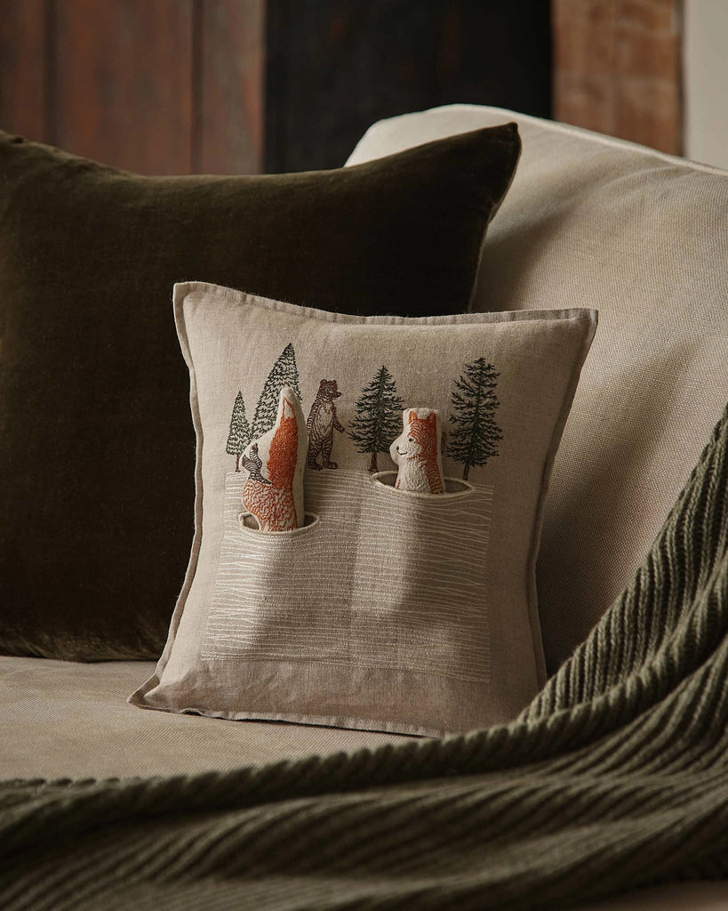 The Coral & Tusk Winter Foxes Pocket Pillow, featuring two foxes in tree holes watching a bear among pine trees, adds a charming winter touch to any beige couch with green accents.