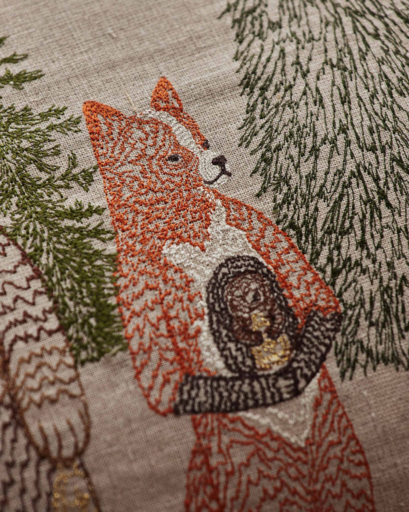 Close-up of a Coral & Tusk Christmas Cheer Lumbar Pillow, a linen lumbar pillow embroidered with a fox holding a small, round object between two pine trees.