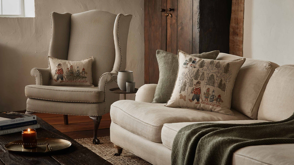 A cozy living room features a beige striped armchair and light sofa with decorative pillows, including the Coral & Tusk Winter Fun Pillow. Holiday décor, a green throw blanket, and a candle on the wooden table create an inviting atmosphere.
