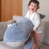 A young child with curly hair and a white top happily holds a large Ride-On Whale Pouf while standing on a beige couch indoors.