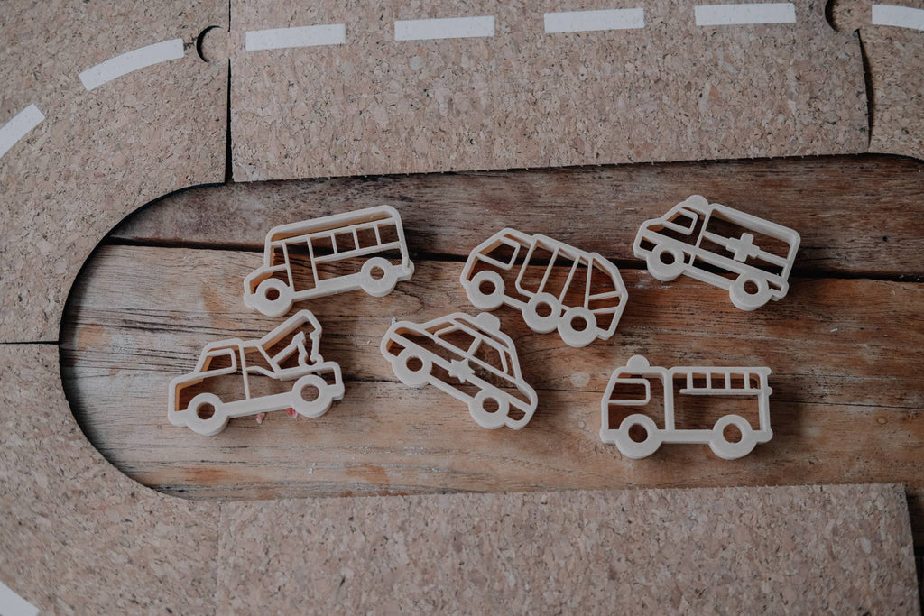 The Service Vehicle Cutter Set - Eco Playdough Tool features wooden cutouts of a bus, ambulance, tow truck, garbage truck, police car, and fire truck arranged on a wooden surface beside a toy road track—perfect for sensory play or transport-themed activities.