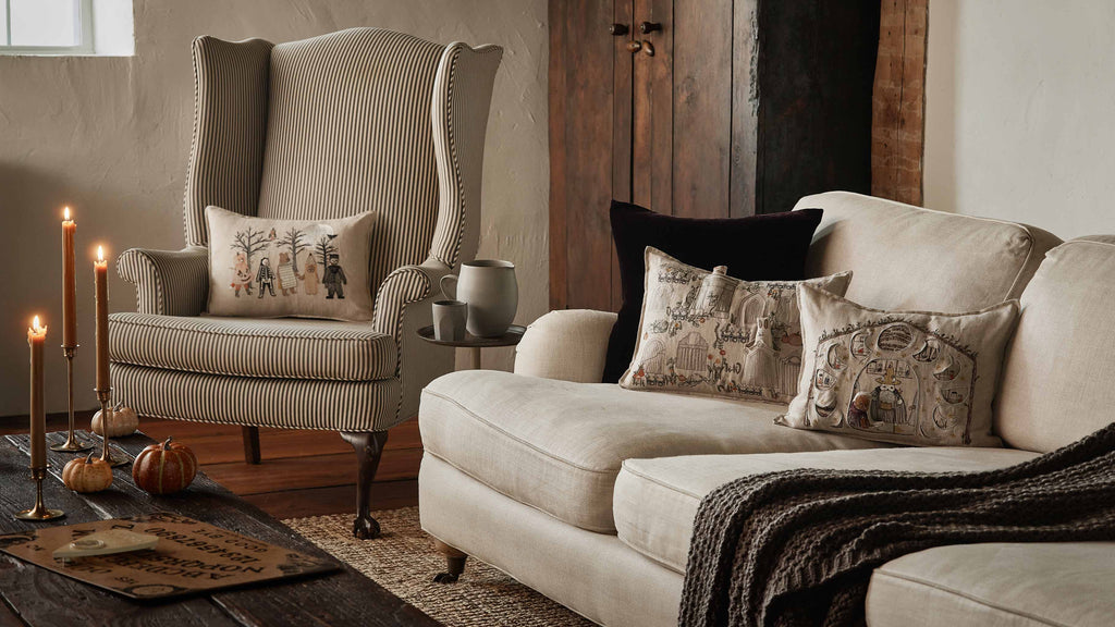 A cozy living room with a striped armchair, beige sofa, Coral & Tusk Full Moon Masquerade Pillow, decorative pumpkins, Ouija board, and lit candles on a wooden coffee table creates a warm autumn atmosphere.