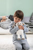 A young child hugs a Steiff, Lita Lamb Plush Animal Toy, 12 Inches, smiling as they kneel on a white carpet with teddy bears and a gray cushion in the background.