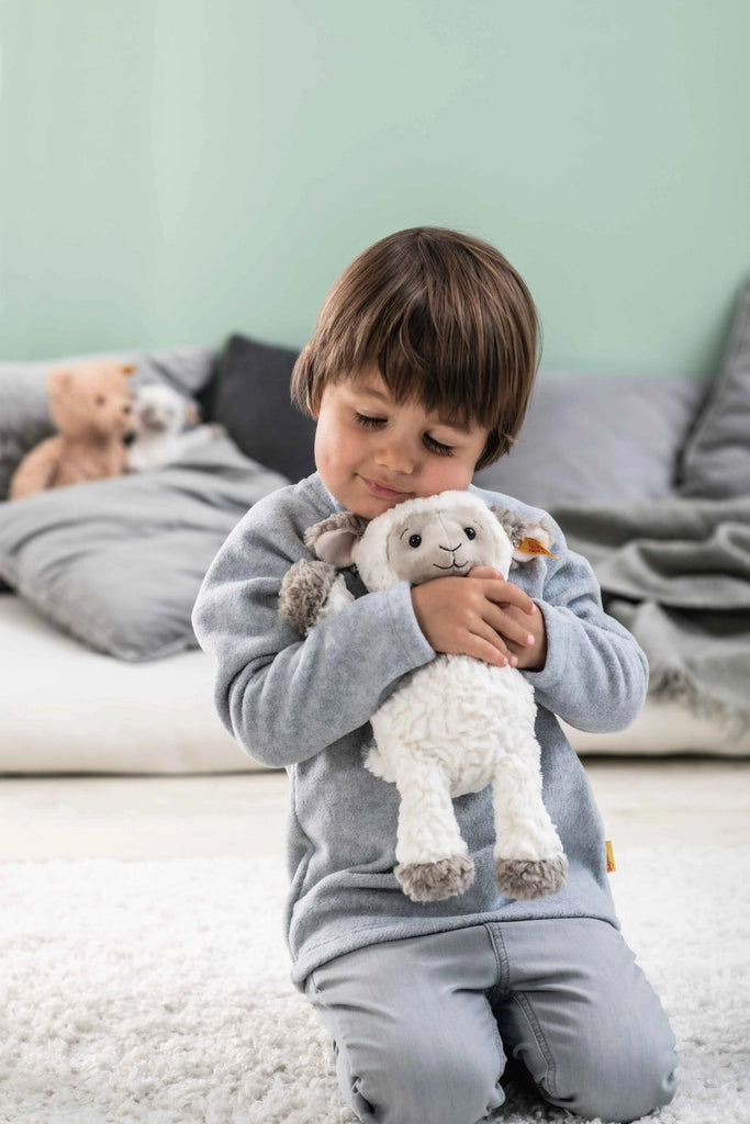A young child hugs a Steiff, Lita Lamb Plush Animal Toy, 12 Inches, smiling as they kneel on a white carpet with teddy bears and a gray cushion in the background.