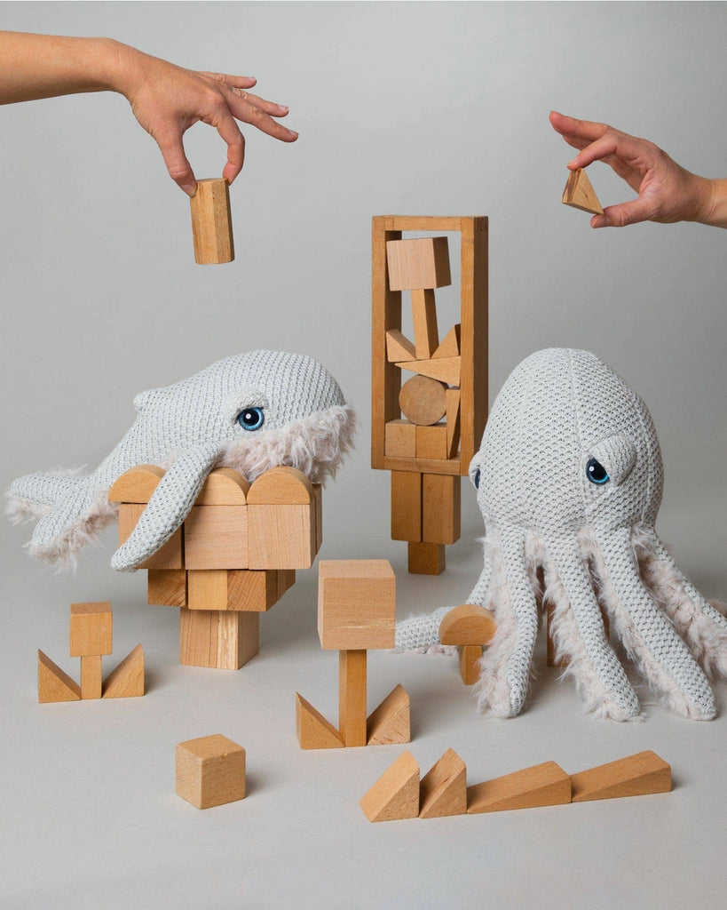 Two hands arrange wooden blocks in front of knit stuffed animals, including a BigStuffed Mini Grandma Whale and an octopus. The toys and blocks are positioned to create different playful structures on a gray background, showcasing the charm of items made in Lithuania.