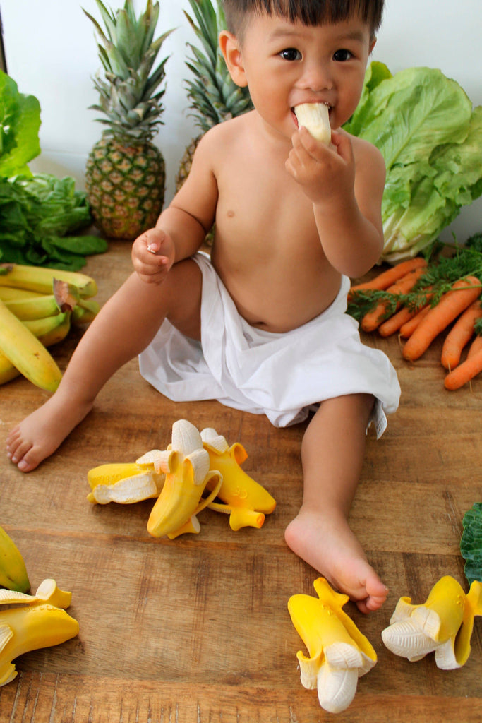 A young child sits on a wooden floor surrounded by bananas, pineapples, lettuce, and carrots, enjoying sensory play with the Ana Banana Teether while eating a banana.