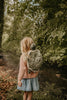 A young child with long blonde hair, in a pink sweater and blue skirt, stands by a forest creek wearing the Children's Backpack - Turtle with adjustable shoulder straps and is facing away from the camera.