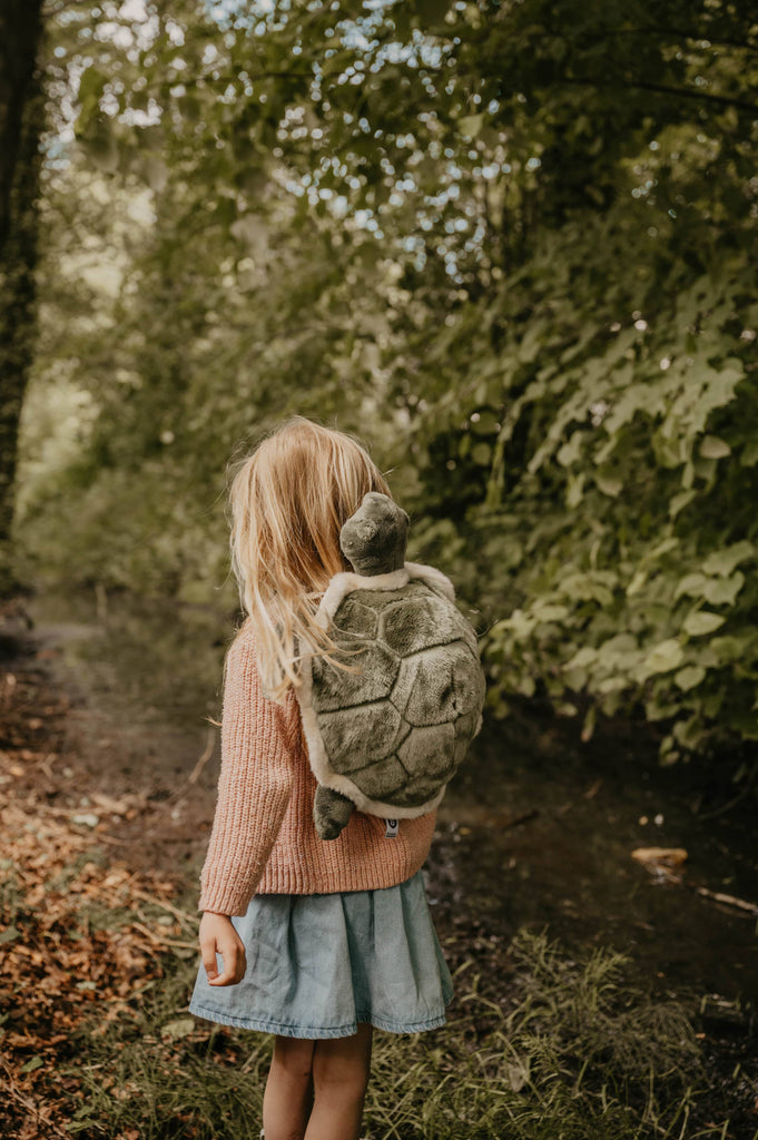A young child with long blonde hair, in a pink sweater and blue skirt, stands by a forest creek wearing the Children's Backpack - Turtle with adjustable shoulder straps and is facing away from the camera.