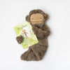 A Slumberkins Bigfoot Snuggler + Intro Book - Self Esteem resembling a Bigfoot snuggler character lying next to a book titled "bigfoot" on a white background. The toy has a friendly face with a slight smile.