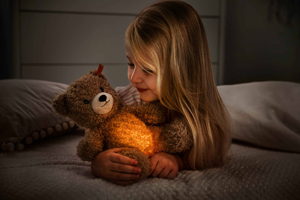 A young girl with long blonde hair lies on her bed, hugging her Light at Night Caspar Teddy Bear Stuffed Plush. The 11-inch bear emits a warm, orange glow from its tummy, creating a cozy atmosphere in the dimly lit room. She smiles softly at the glowing bear that charges easily via USB.