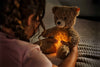 A child in a pink shirt, with braided hair, holds the 11-inch Light at Night Caspar Teddy Bear Plush, which emits a soft light from its belly. The dimly lit room creates a cozy atmosphere as the bear, USB rechargeable, captures the child's attention.