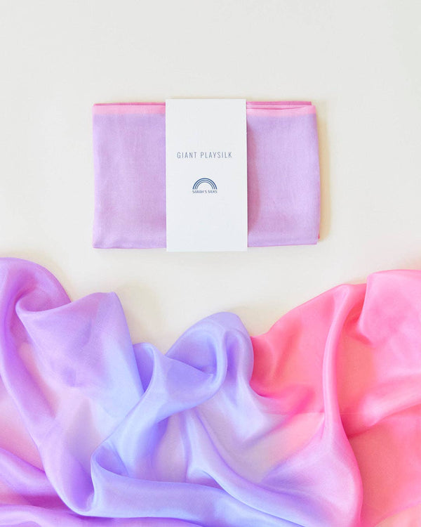 A folded Sarah's Silk Giant Blossom Playsilk in pastel purple is displayed above a similar playsilk draped in a gradient of pink, purple, and blue, ideal for open-ended play. The label features a simple rainbow logo against a soft cream background.