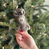 A hand with red polished nails holds a small Donkey Finger Puppet, crafted from 100% wool and featuring a brown color. In the background, there is a Christmas tree. This charming finger puppet is handmade in Nepal.