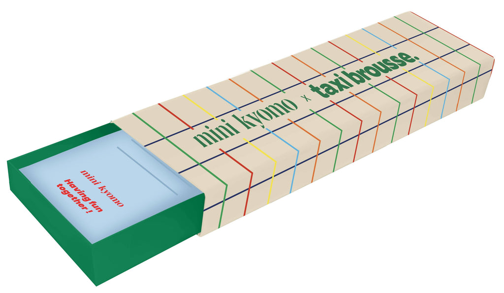 A rectangular box with a colorful grid and “Mini Kyomo Watch x Taxi Brousse - Color Notebook” text is partially open, showing a light blue interior with red text, reflecting the product’s vintage style.