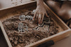A child’s hand uses the Construction Cutter Set - Eco Playdough Tool to shape construction vehicle-themed pieces in a tray filled with brown kinetic sand, creating an engaging sensory play activity. The child’s knees are visible in the background, emphasizing the hands-on fun inspired by the Eco Playdough tool.