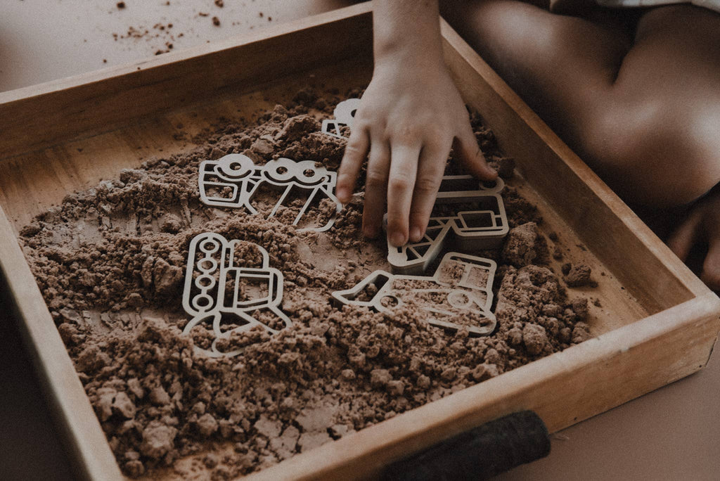 A child’s hand uses the Construction Cutter Set - Eco Playdough Tool to shape construction vehicle-themed pieces in a tray filled with brown kinetic sand, creating an engaging sensory play activity. The child’s knees are visible in the background, emphasizing the hands-on fun inspired by the Eco Playdough tool.