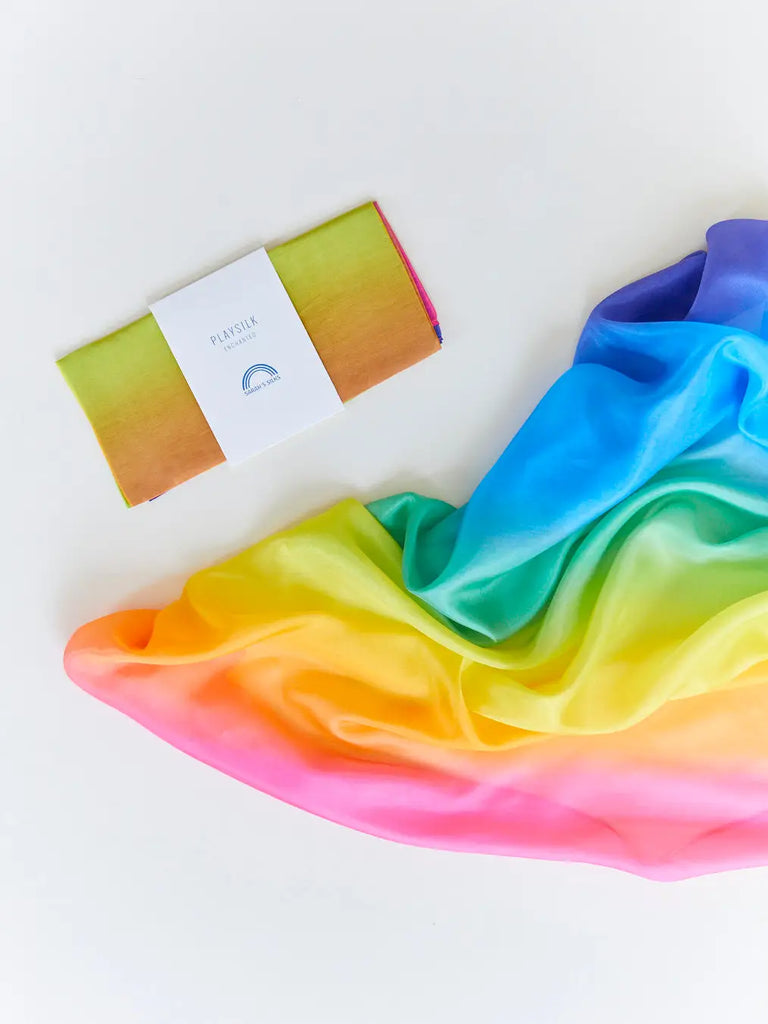 A bar of soap with a rainbow gradient resting on a flowing, silky Sarah's Silk Enchanted Playsilk - Rainbow featuring vibrant rainbow colors against a white background.