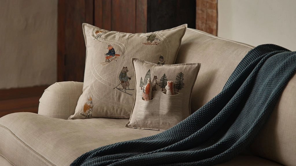 The Coral & Tusk Winter Foxes Pocket Pillow with embroidered fox and tree designs sits on a beige sofa beside a dark green knitted throw, creating a cozy and inviting atmosphere.
