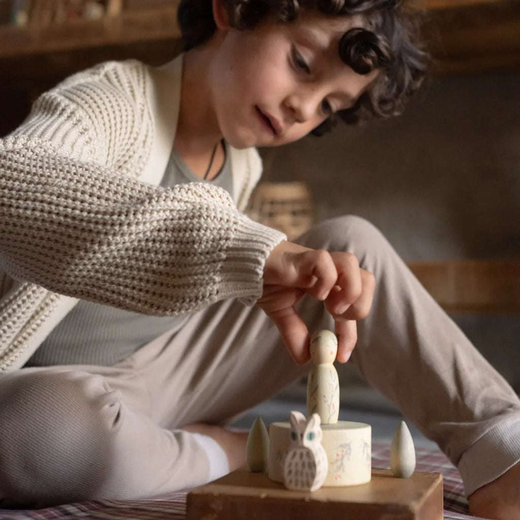 A young child with curly hair and a knit sweater carefully places an Ostheimer figure from the Hello Winter Set (Grapat X Ostheimer Limited Edition) onto a spinning top on the table.