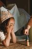 A young child wearing a floral hat smiles on the floor during Quiet Time, watching another child hold a Grapat Quiet Time Advent Calendar 2025 figure, with a cozy blanket fort in the background.
