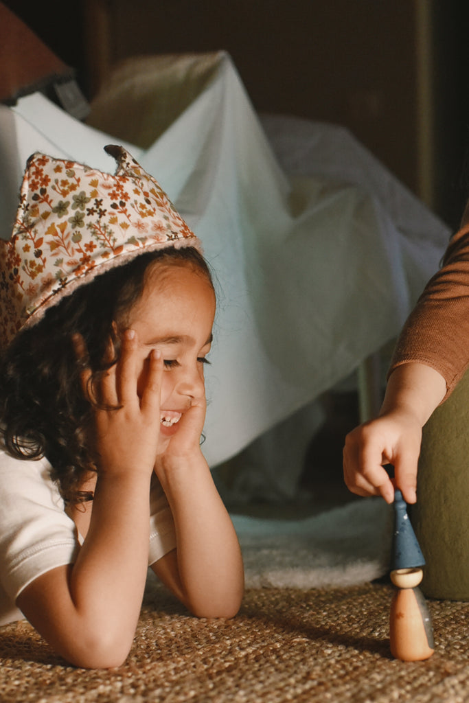 A young child wearing a floral hat smiles on the floor during Quiet Time, watching another child hold a Grapat Quiet Time Advent Calendar 2025 figure, with a cozy blanket fort in the background.