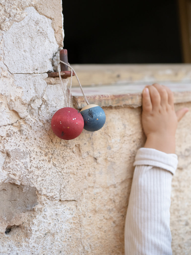 A child's hand in a long-sleeved shirt reaches toward a textured stone wall, where two Grapat Ornament Set - Wish Balls, one red and one blue, hang by strings from a rusty hook.