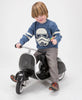 A young child with a joyful expression rides a PRIMO Ride On Kids Toy Classic, wearing a blue sweater with a classic Star Wars stormtrooper design, gray pants, and colorful shoes, on a white background.