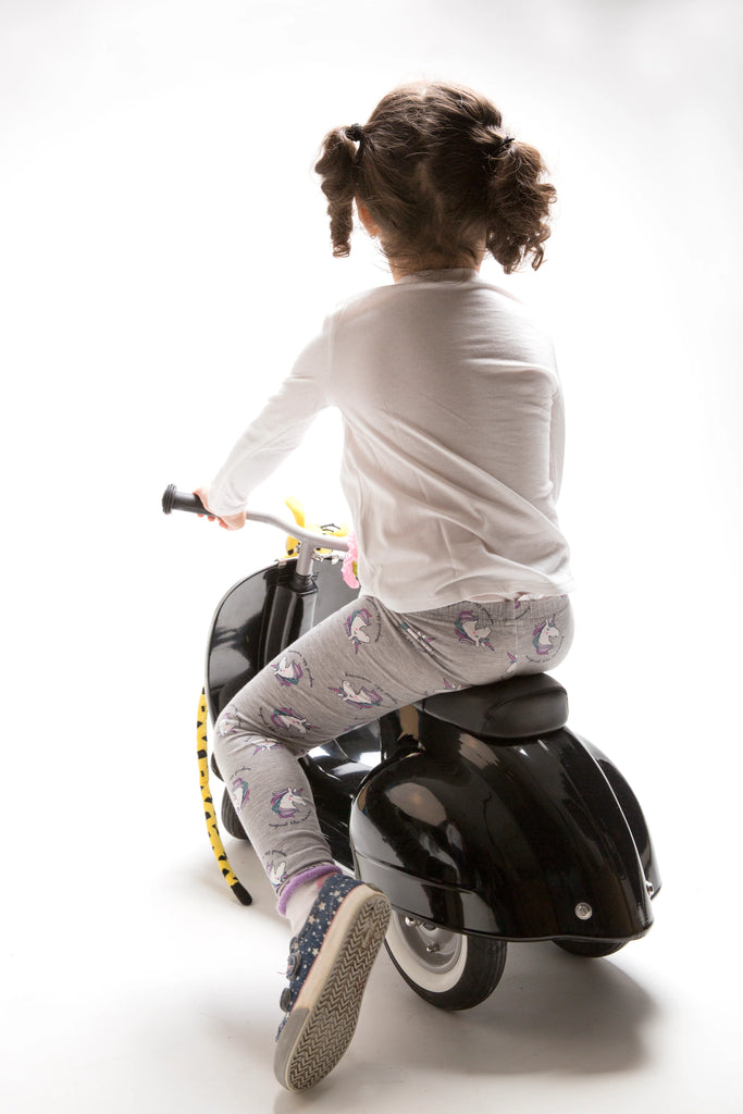 A young girl with pigtails, wearing a white long-sleeve shirt and patterned leggings, rides on a PRIMO Ride On Kids Toy Classic against a white background. She is seen from the back.