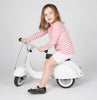 A young girl with light brown hair, wearing a pink and white striped shirt and shorts, rides a small white PRIMO Ride On Kids Toy Classic scooter, looking at the camera with a playful expression.