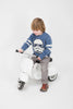 A young child with shoulder-length hair, wearing a blue sweater featuring a stormtrooper design, rides a white PRIMO Ride On Kids Toy Classic scooter against a plain background.