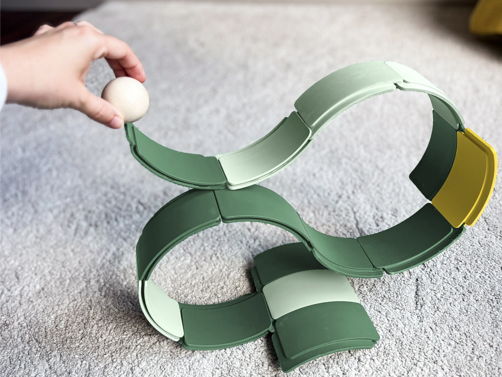 A hand holds a wooden ball above the WaytoPlay: Arches - Starter Set, a looping green and yellow sustainable toy marble run, set up on a light carpeted floor.