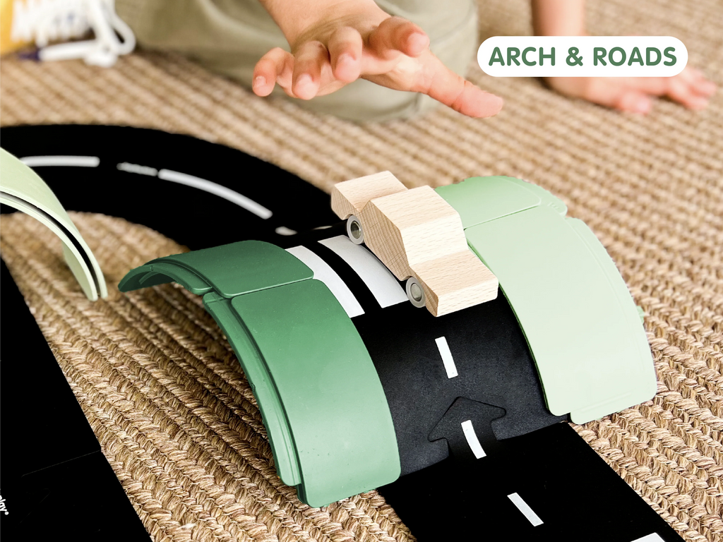 A child’s hand reaches toward a wooden toy car on a curved track with green arches, placed on a textured rug. A sign in the corner reads “WaytoPlay: Arches - Starter Set.” This creative construction toy encourages imaginative play and highlights the beauty of arches made from sustainable materials.