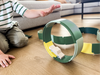 A child in a striped shirt plays on a wooden floor, launching a small wooden toy car through the green and yellow circular track of the WaytoPlay: Arches - Creator Set, with more creative construction pieces from the set scattered in the background.