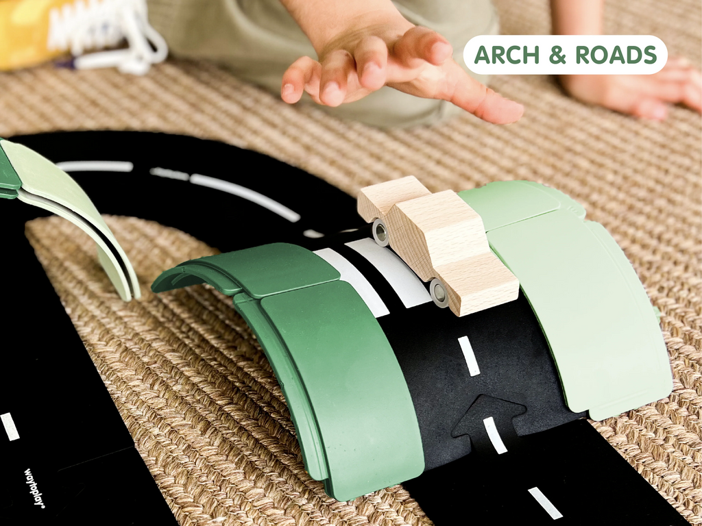 A child’s hand reaches toward a wooden toy car on a curved, modular road track set atop a woven rug—the perfect setting for imaginative play with the WaytoPlay: Arches - Creator Set. “ARCH & ROADS” appears in a green-bordered box in the top right corner.