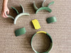 A child’s hand arranges green and yellow plastic curved toy pieces from the WaytoPlay: Arches - Builder Set on a woven beige rug, forming abstract shapes.