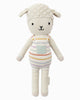 Cuddle + Kind Avery the Lamb is a hand-knit, cream-colored doll made from 100% cotton yarn, featuring a curly head, floppy ears, and pastel striped overalls with a light blue pocket.