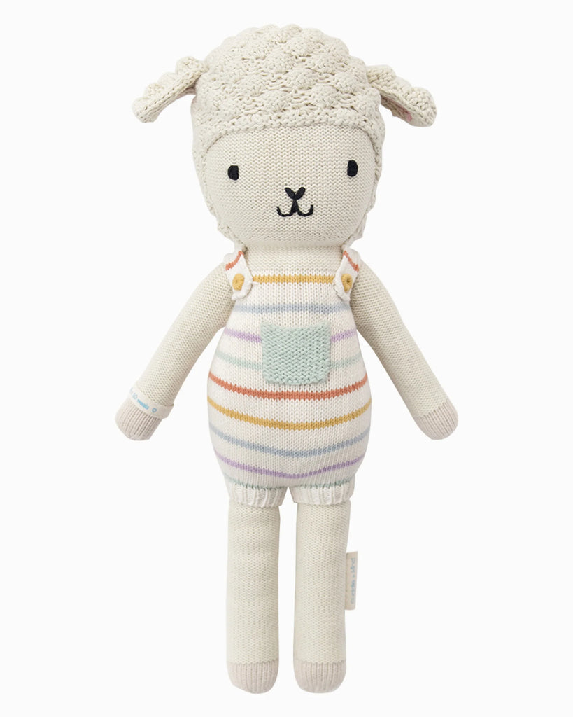 Cuddle + Kind Avery the Lamb is a hand-knit, cream-colored doll made from 100% cotton yarn, featuring a curly head, floppy ears, and pastel striped overalls with a light blue pocket.