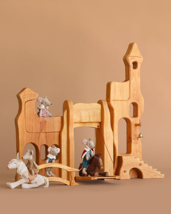 Four Maileg Royal Family mice enjoy the Ostheimer Wooden Kingdom—one rides a unicorn, one swings on a horse, and two perch on towers. A bell hangs on the right, set against a light brown background.