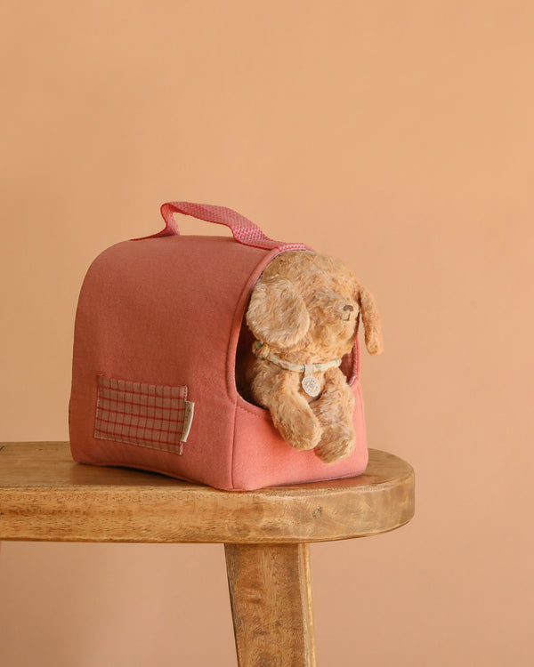 The Maileg Puppy in Coral Pet Carrier - Dusty Powder sits on a wooden stool. The soft pink carrier with a handle holds the plush puppy, set against a plain beige background.