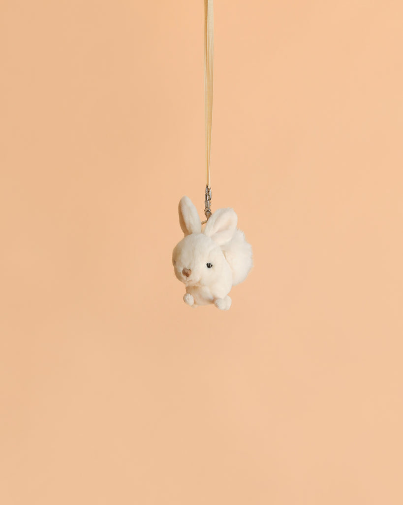 The Bukowski - Zeus Bunny Bag Charm, a small white plush bunny keychain, is suspended by a ribbon against a plain peach-colored background.